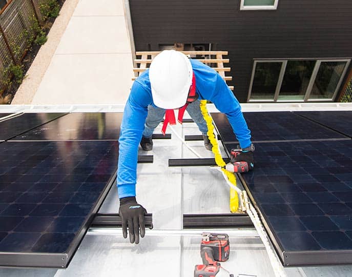 Solar technician wearing a royal blue shirt and white hard hat, on ladder installing solar panels, shot looking down to the ground.