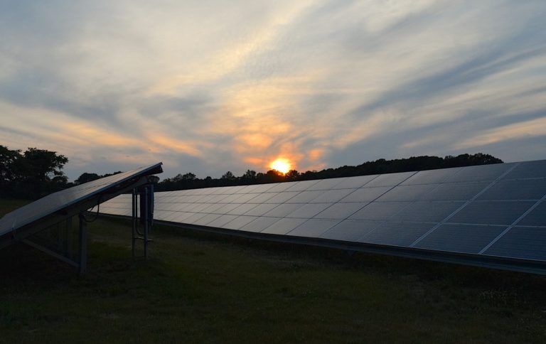 A solar panel array installed in a field with a view of the sun setting in the distance.