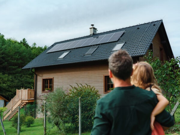 Rear view of dad holding her little girl in arms and looking at their house with installed solar panels. Alternative energy, saving resources and sustainable lifestyle concept.