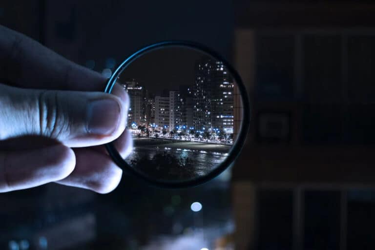 Close-up of a hand holding a magnifying glass pointed at a city skyline at night.
