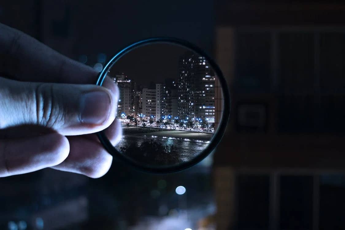 Close-up of a hand holding a magnifying glass pointed at a city skyline at night.