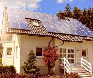 A small white house with the sun shining down upon its rooftop solar panel installation