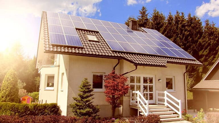 A small white house with the sun shining down upon its rooftop solar panel installation