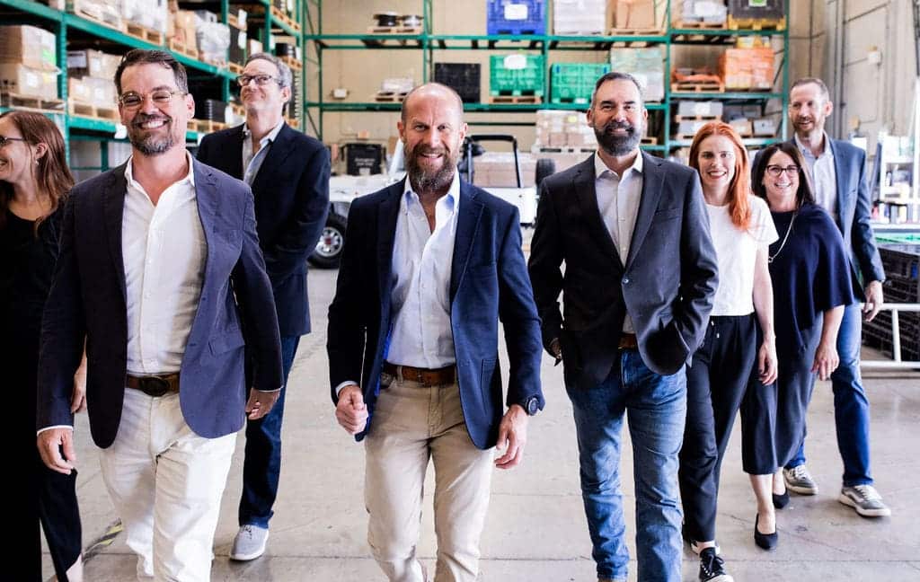 8 people in professional attire walk in a solar panel warehouse, smiling and walking towards the camera