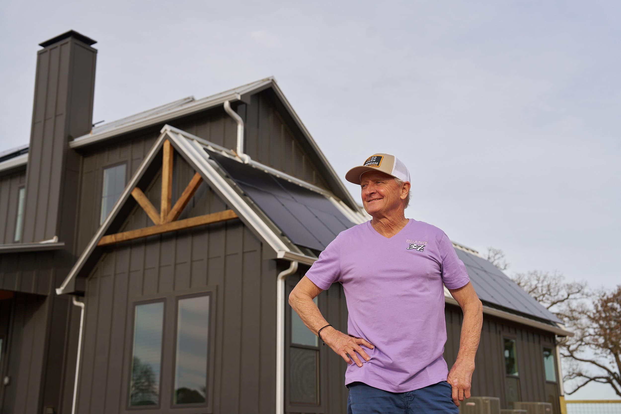 Smiling man in purple t-shirt standing in front of modern ranch home with solar panels on roof.